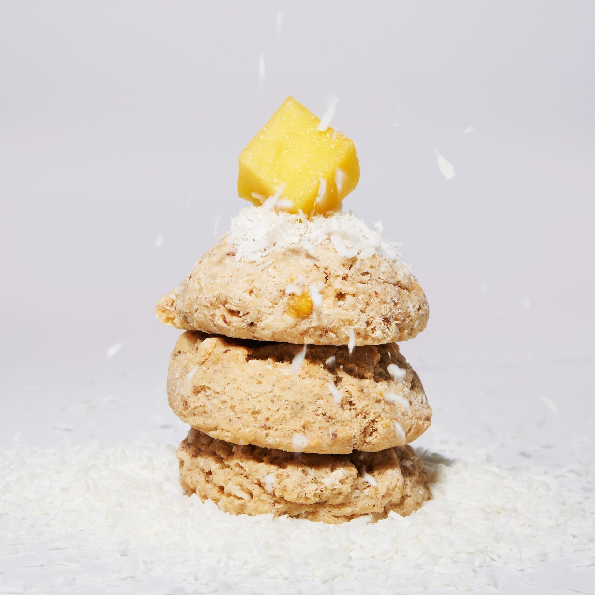 Three Mango Coconut Lime Cookies with coconut falling from the sky and a piece of mango on top in front of a grey background.. 