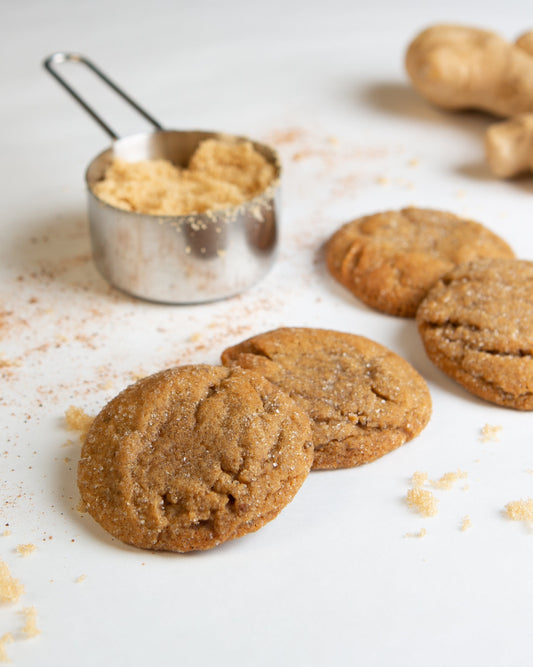 Four ginger molasses cookies on a white background with a cup of brown sugar and a thumb of ginger in the background.